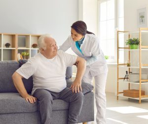 Nurse helping senior man to stand up from sofa in modern hospital or geriatric rehabilitation center